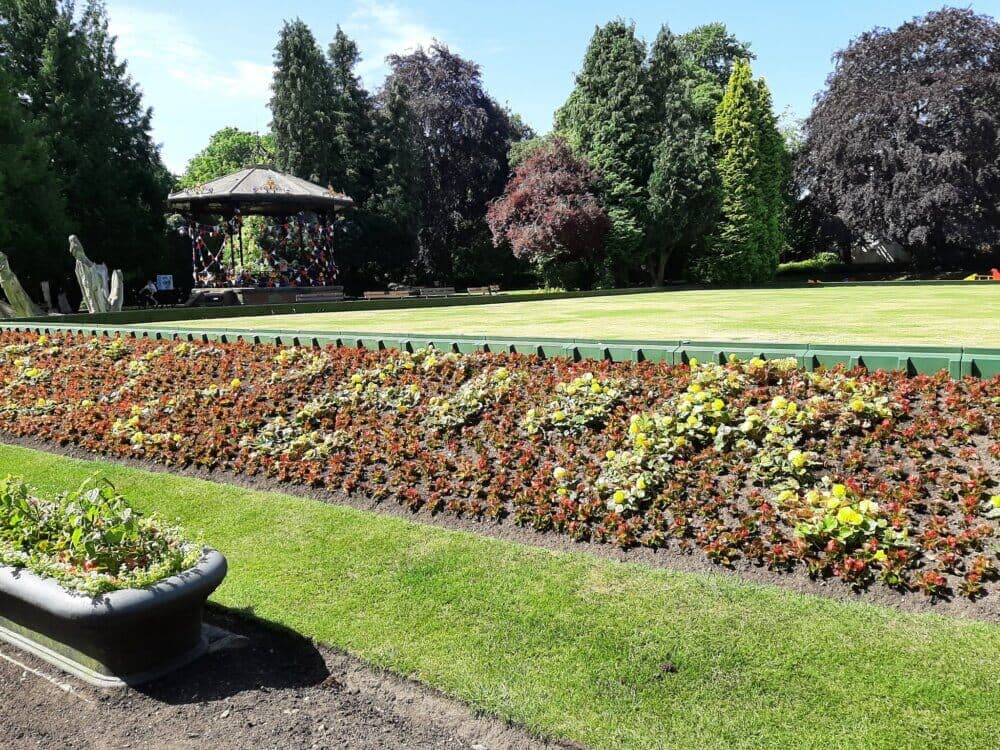 A vibrant garden with flower beds, a grassy lawn, and a bandstand surrounded by tall trees on a sunny day. - Home Instead
