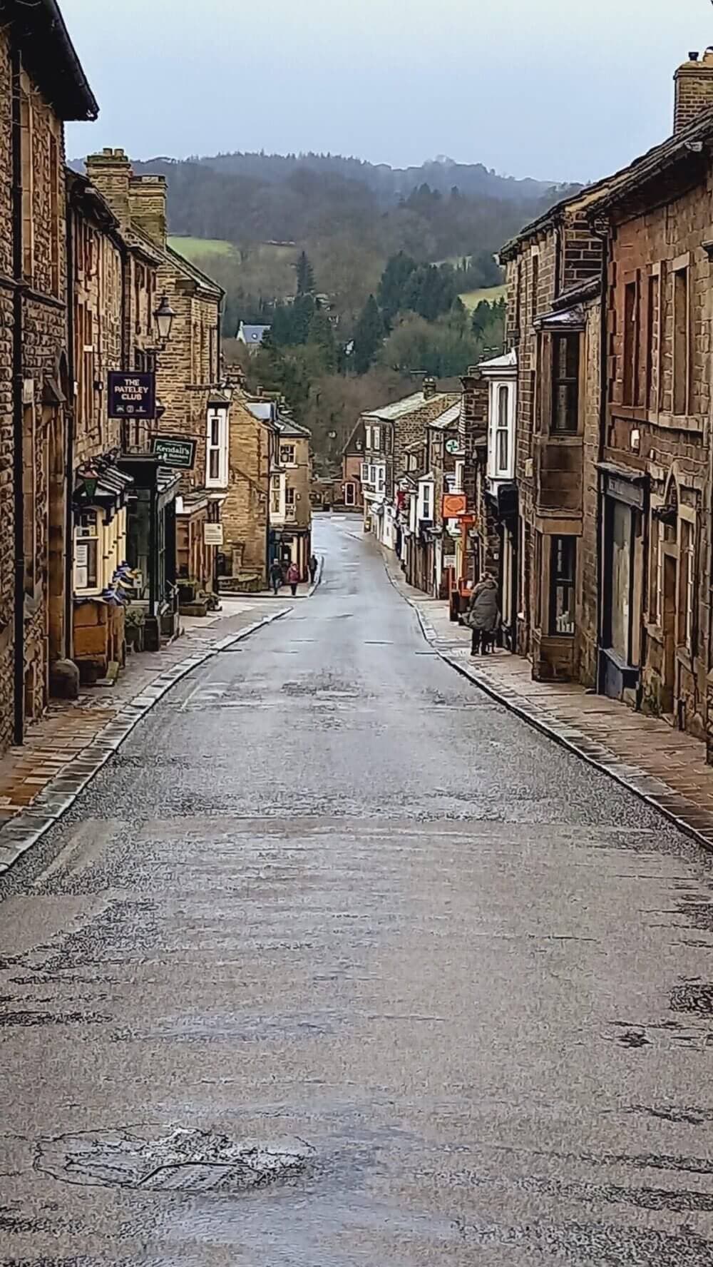 A narrow, empty street lined with historic buildings on a rainy day, leading towards a forested hillside. - Home Instead
