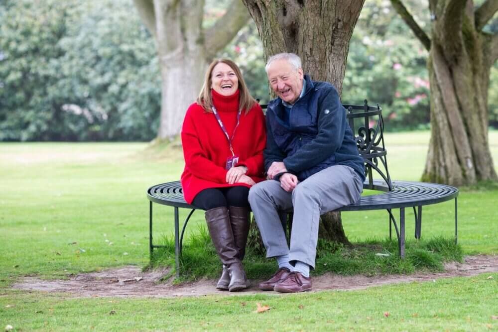 Two people sit and laugh on a circular bench under a tree in a green park, wearing casual clothes and jackets. - Home Instead