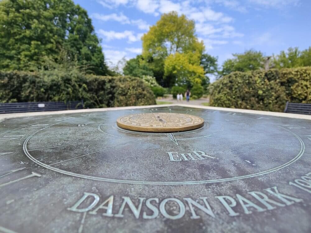 Close-up of sundial reading "Danson Park" with a background of trees, bushes, and a blue sky. - Home Instead