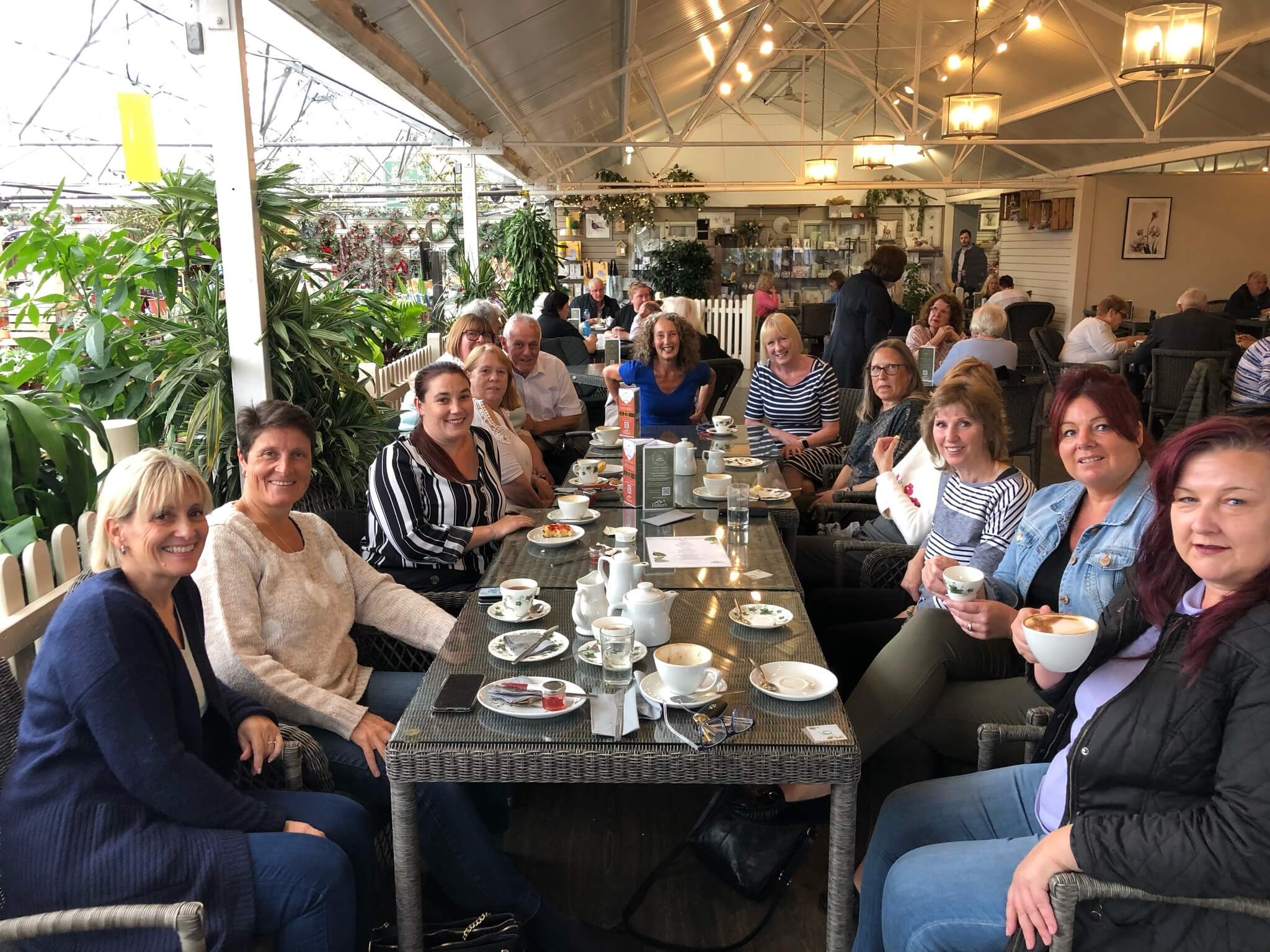 A group of people sitting around a large table in a bright, indoor café setting, smiling and enjoying drinks. - Home Instead
