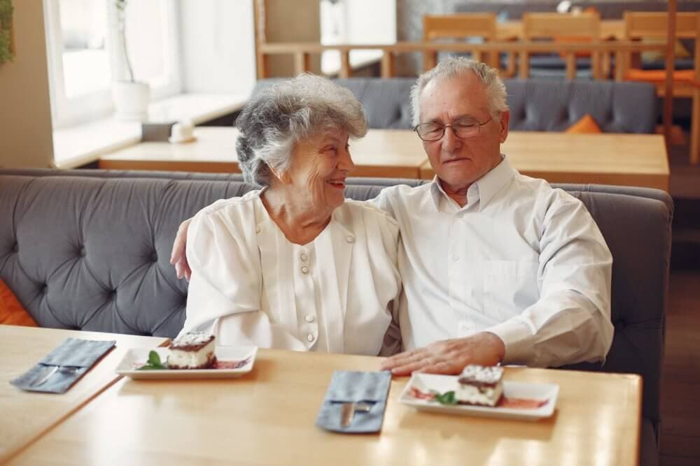 Elderly couple in white clothes sitting closely at a table with dessert, smiling and enjoying each other's company. - Home Instead