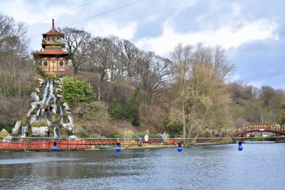 A pagoda with a waterfall sits beside a lake surrounded by trees and a bridge in a serene park setting. - Home Instead