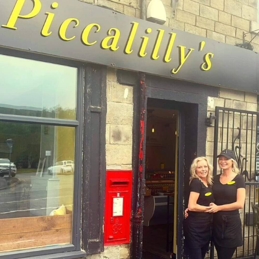 Two women in black uniforms stand smiling outside a storefront with a sign that reads "Piccalilly's. - Home Instead