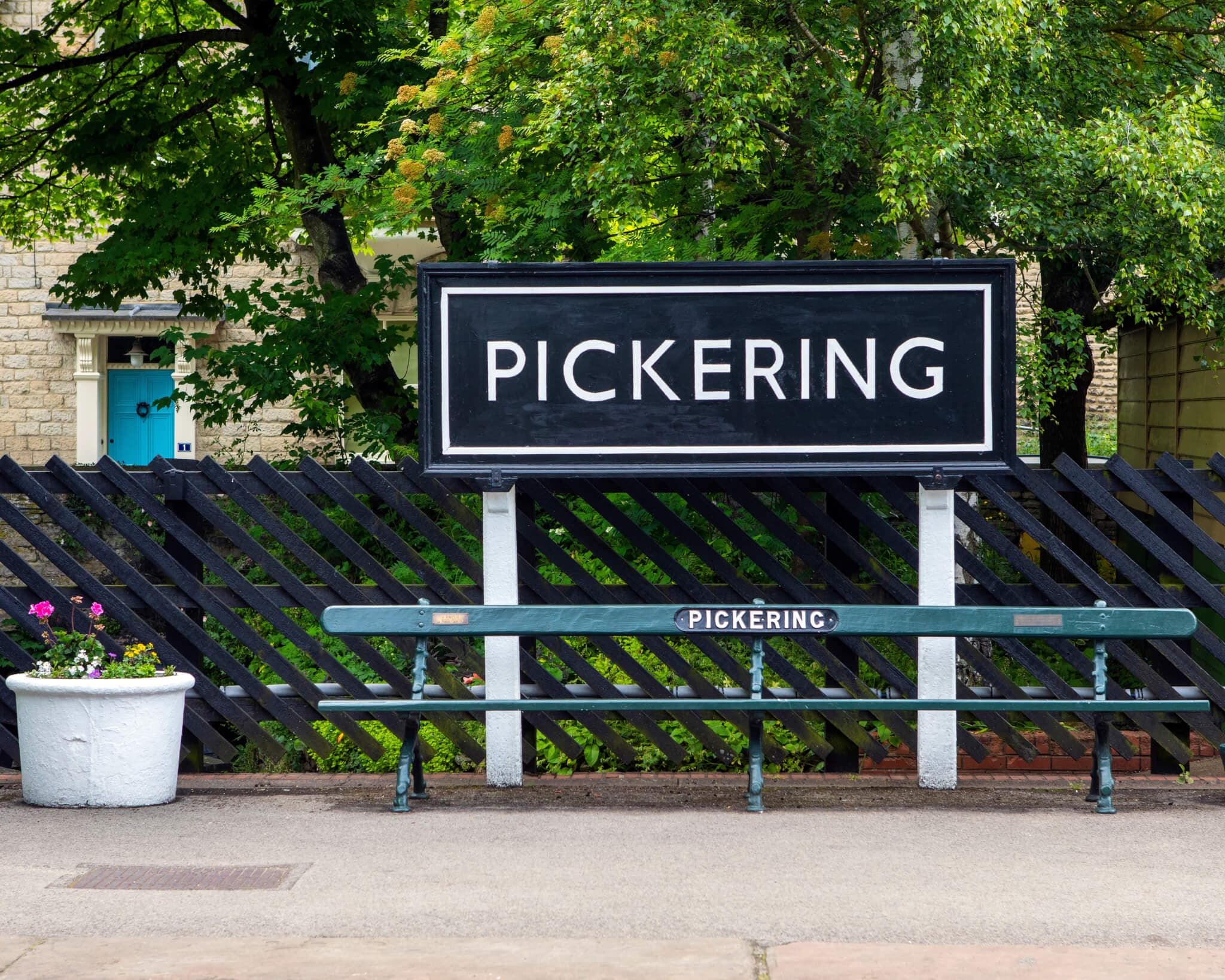 A black sign with white letters reading "Pickering" in front of a bench and black wooden fence. Trees are in the background. - Home Instead