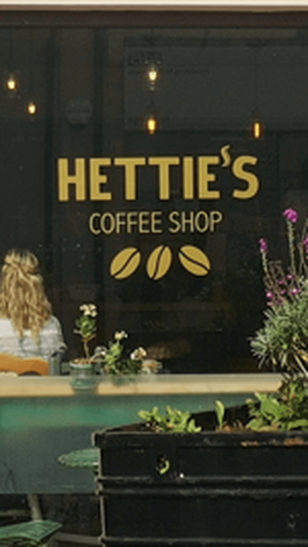 Hettie's Coffee Shop window with logo and plants in front, and a woman with blonde hair seated inside. - Home Instead
