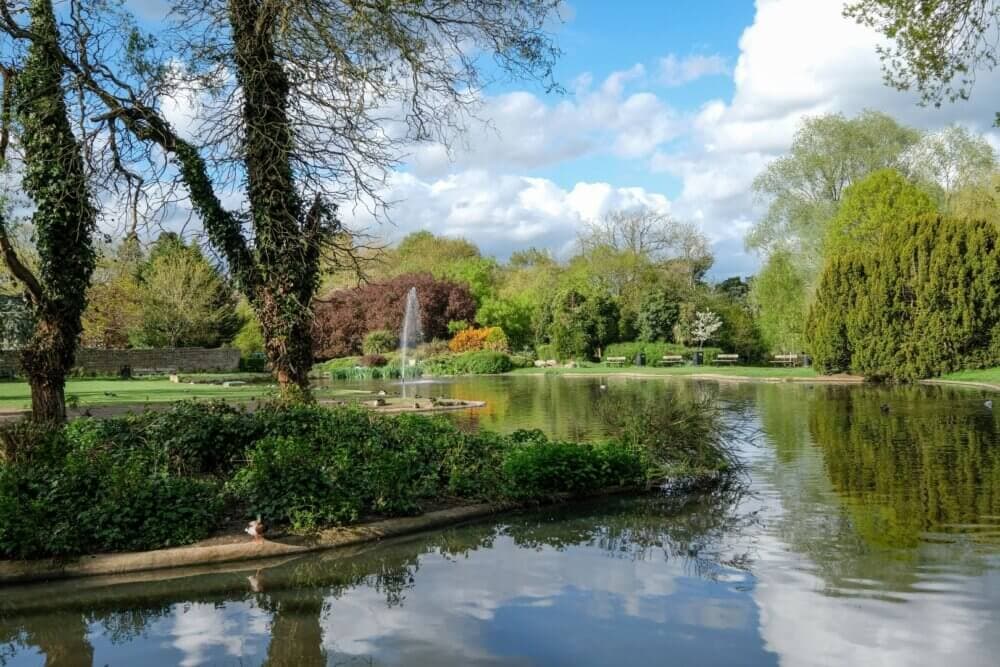 A serene park scene with a pond, fountain, lush greenery, trees, and a partly cloudy sky. - Home Instead