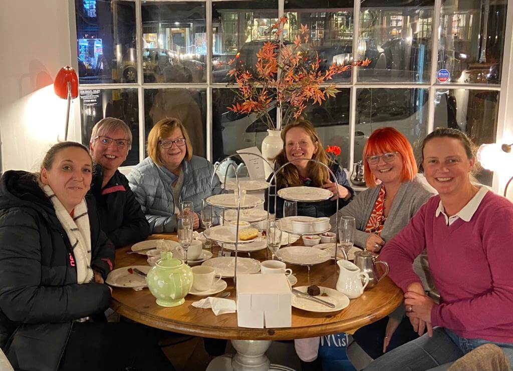 A group of six people smiling around a table in a café with empty plates and a window in the background at night. - Home Instead