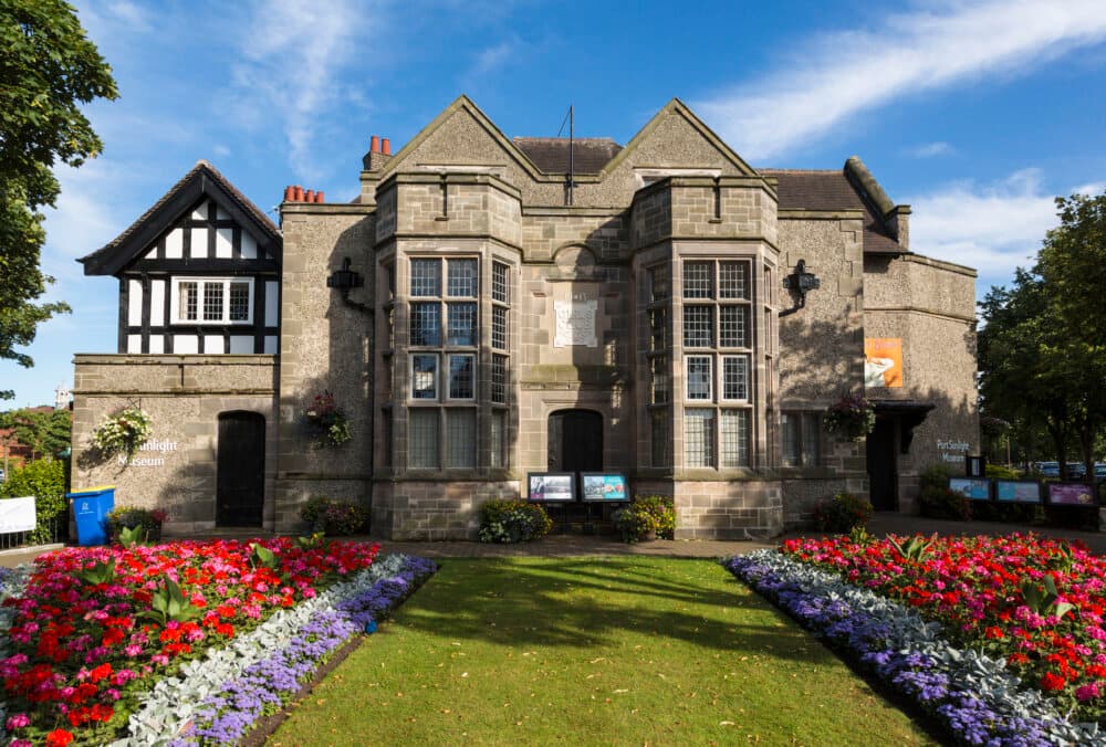Stone building with manicured gardens and colorful flowers in front, under a clear blue sky with scattered clouds. - Home Instead