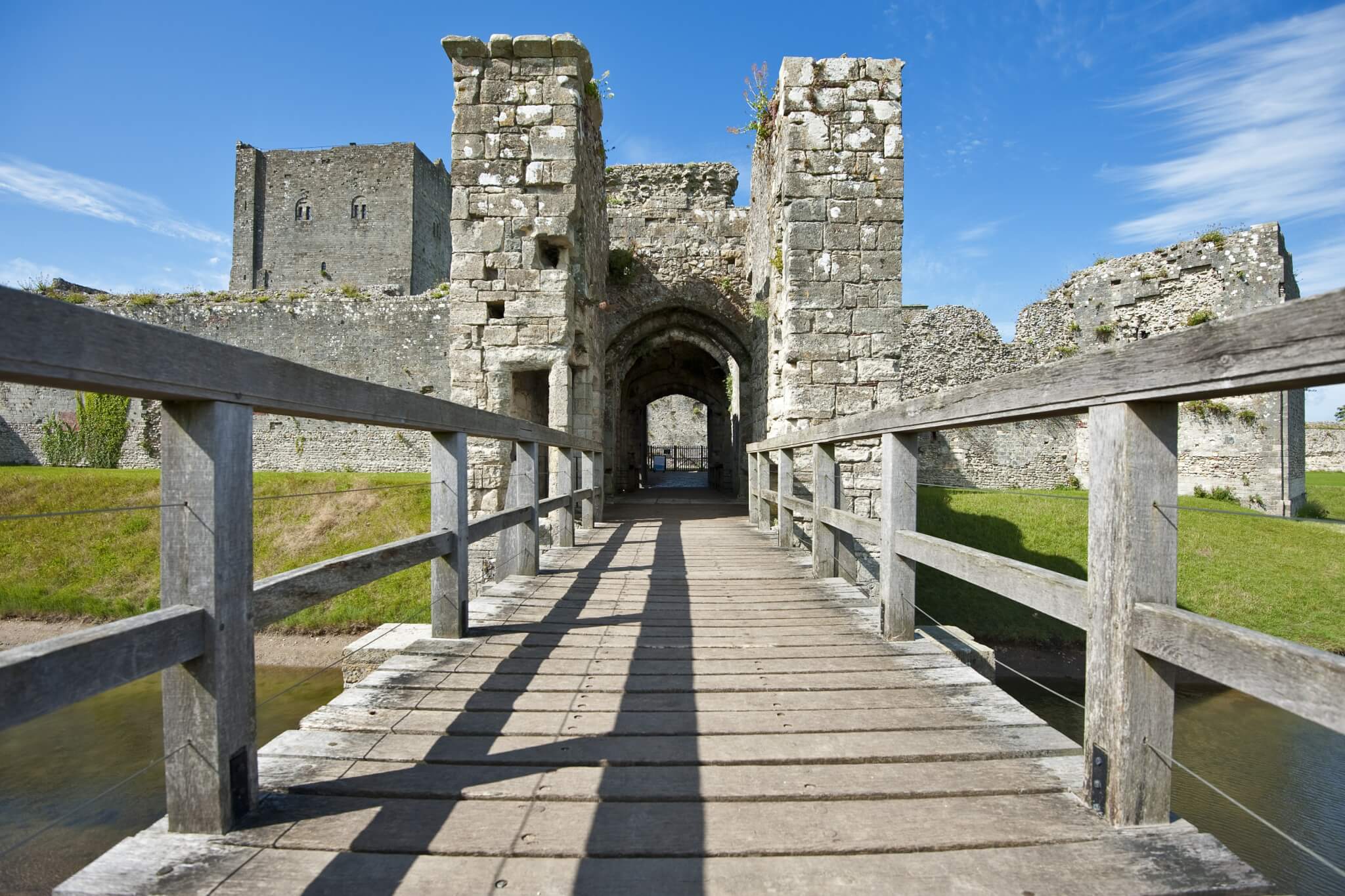 Medieval stone castle ruins with a wooden bridge leading to the arched entrance, surrounded by grass and water. - Home Instead