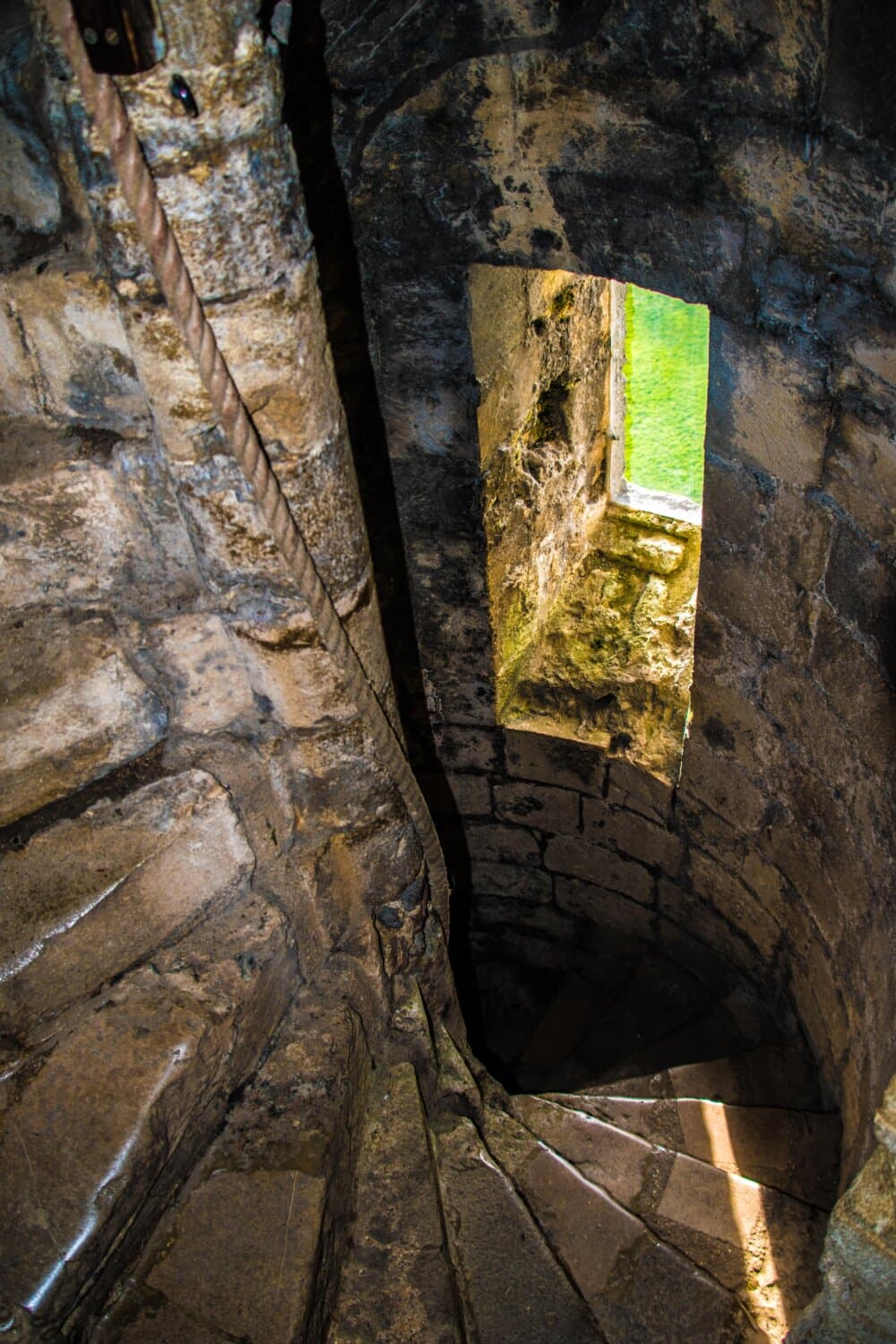 Stone spiral staircase leading downwards with a small window view of green water. - Home Instead