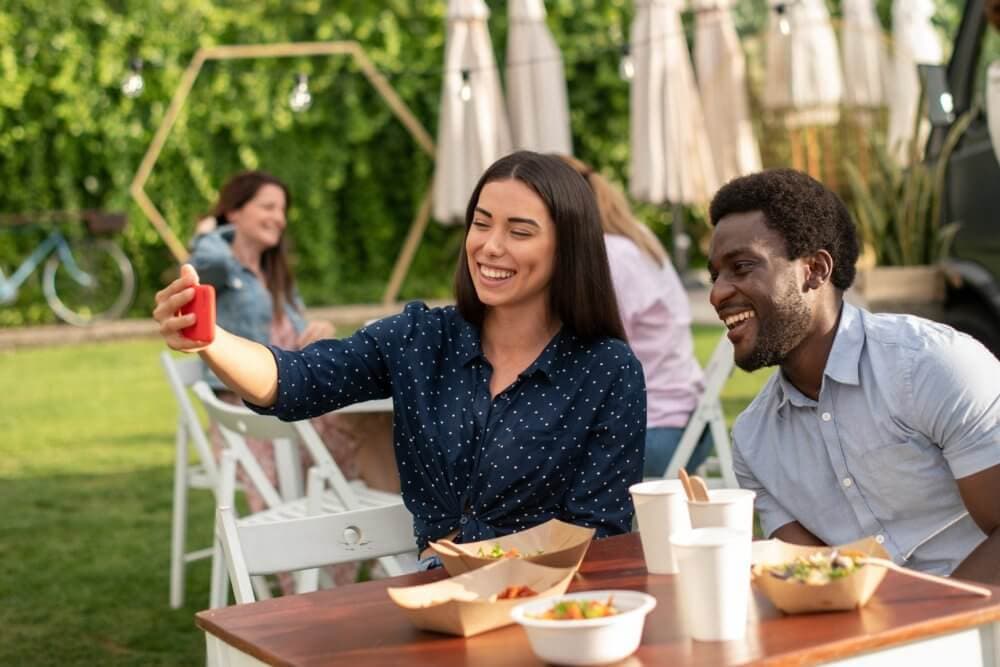 Two people sitting at an outdoor table, smiling and taking a selfie with a smartphone. Food and drinks on the table. - Home Instead