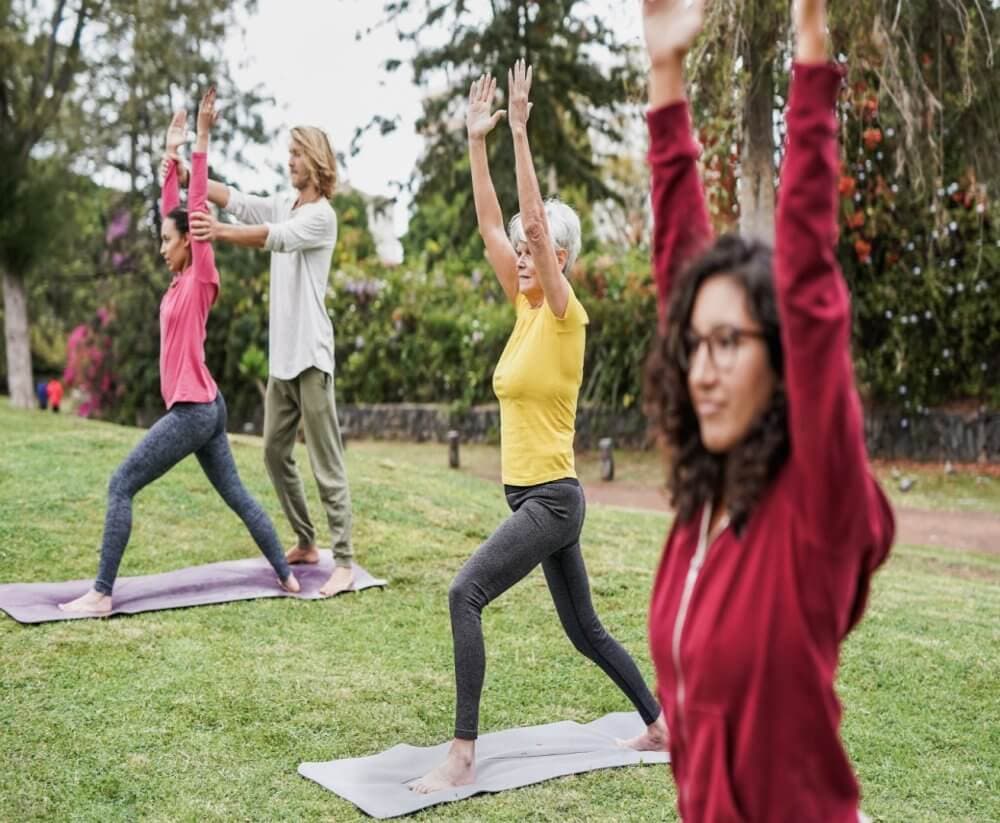 A group of people practicing yoga outdoors on mats, guided by an instructor. Trees and greenery are in the background. - Home Instead
