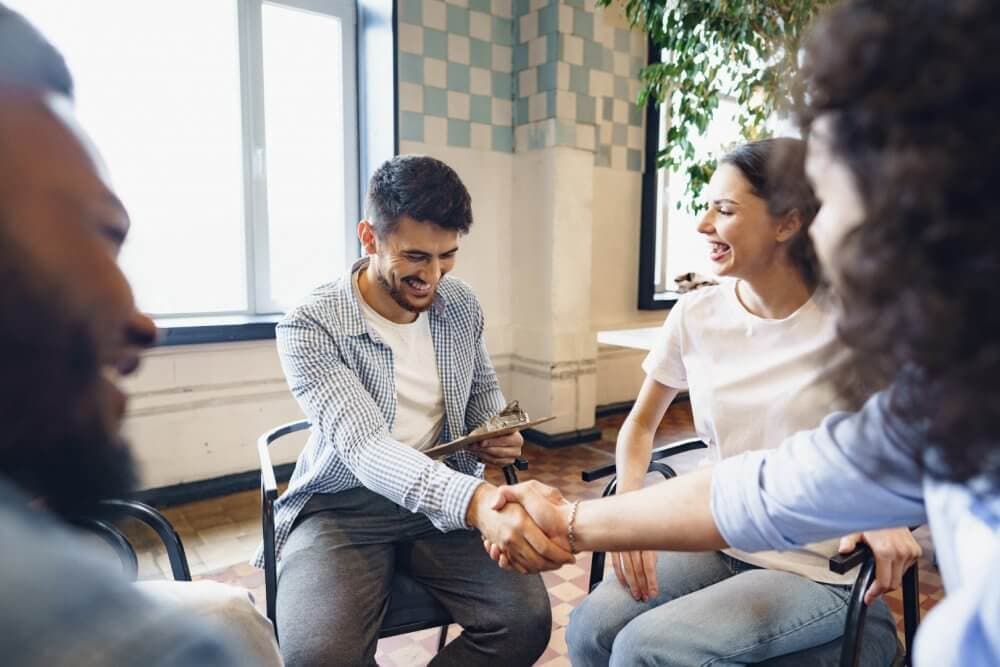 Group of people sitting in a circle indoors, two of them shaking hands and smiling, while others look on cheerfully. - Home Instead