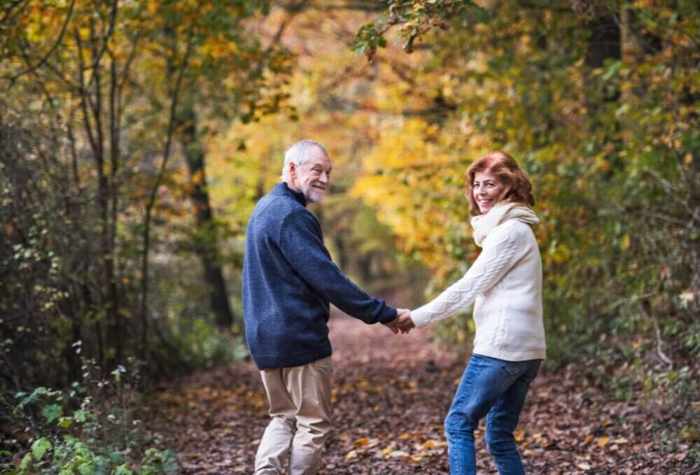 A senior couple holds hands while walking on a forest path surrounded by autumn foliage. - Home Instead