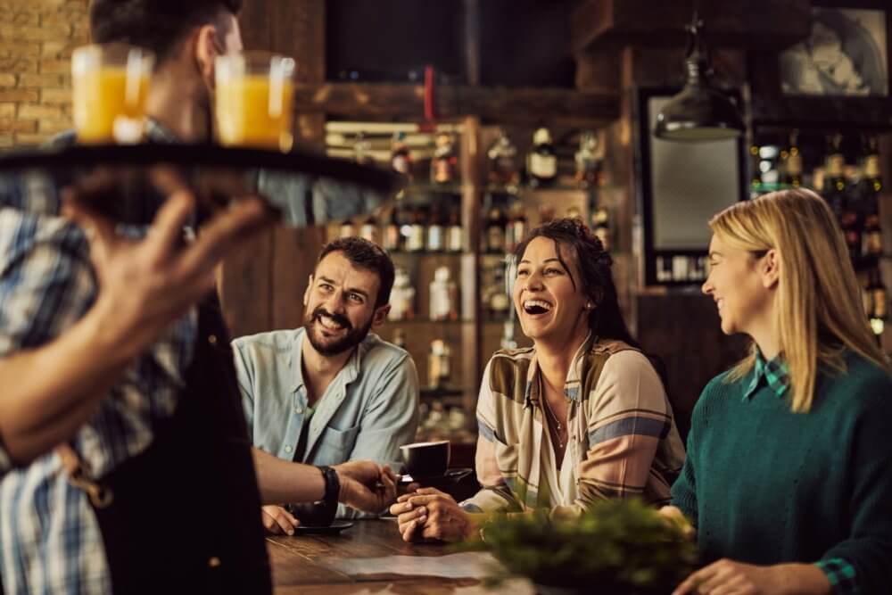 Three people smile and laugh at a bar as a waiter serves drinks. - Home Instead