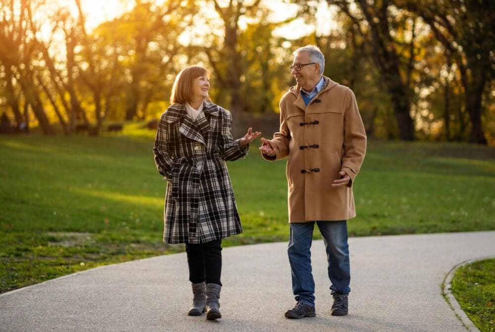 An older couple walks along a park path, talking and smiling, with trees and sunlight in the background. - Home Instead