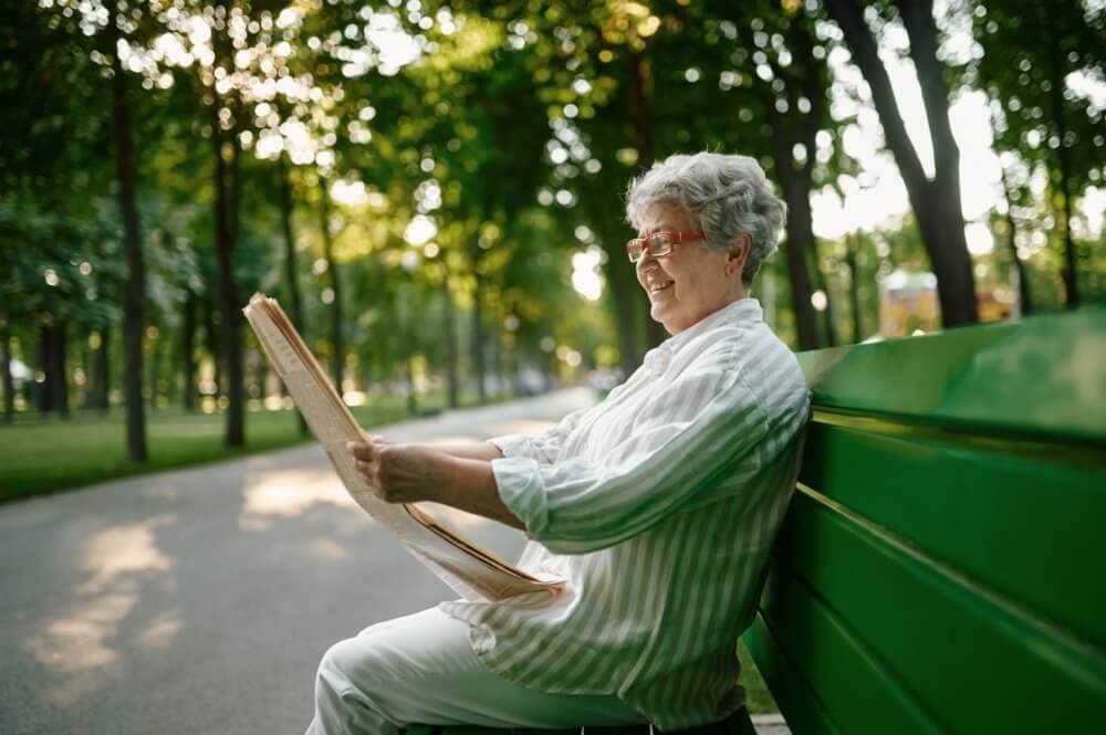 Elderly person with gray hair and glasses smiles while reading a newspaper on a green bench in a park. - Home Instead