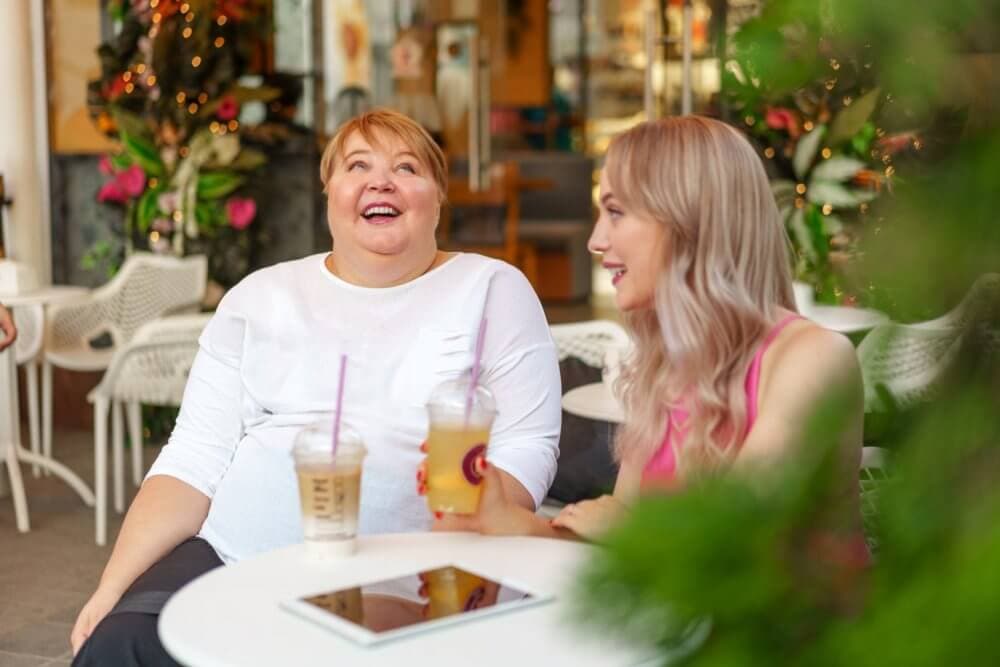 Two women sitting at an outdoor cafe, laughing and holding iced drinks with a tablet on the table in front of them. - Home Instead
