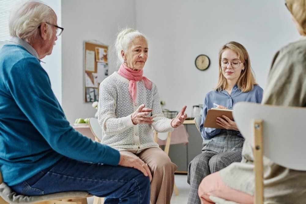 An elderly woman speaks to a group in a support meeting, while a young woman takes notes on a clipboard. - Home Instead