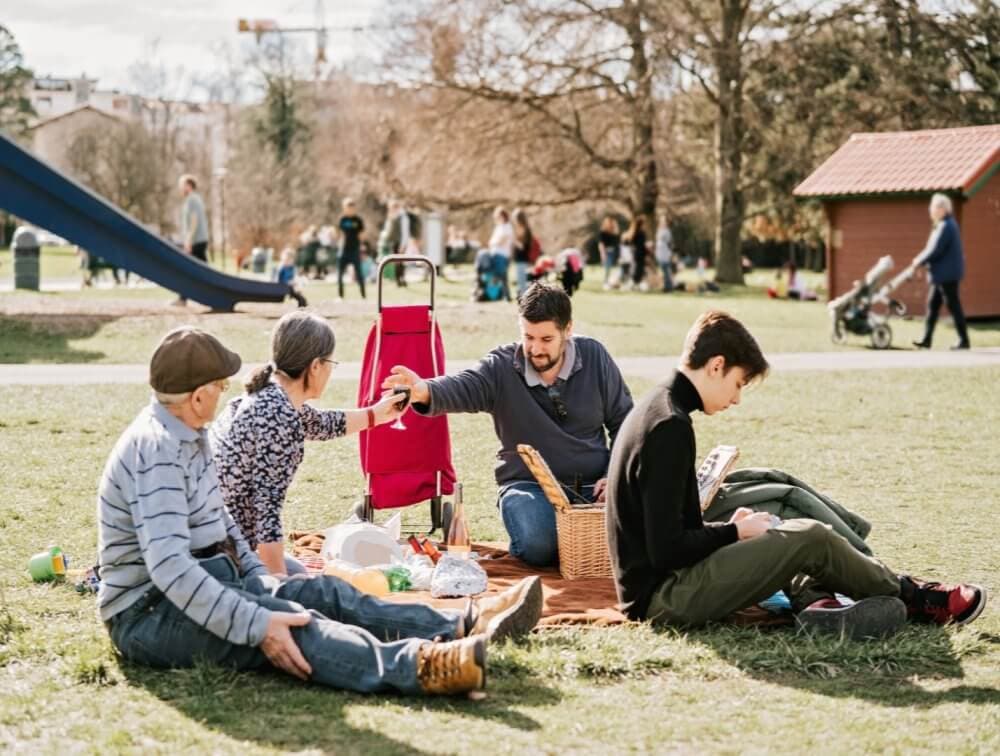 A family enjoys a picnic in a park. They sit on a blanket with food, surrounded by greenery and other park visitors. - Home Instead Southampton