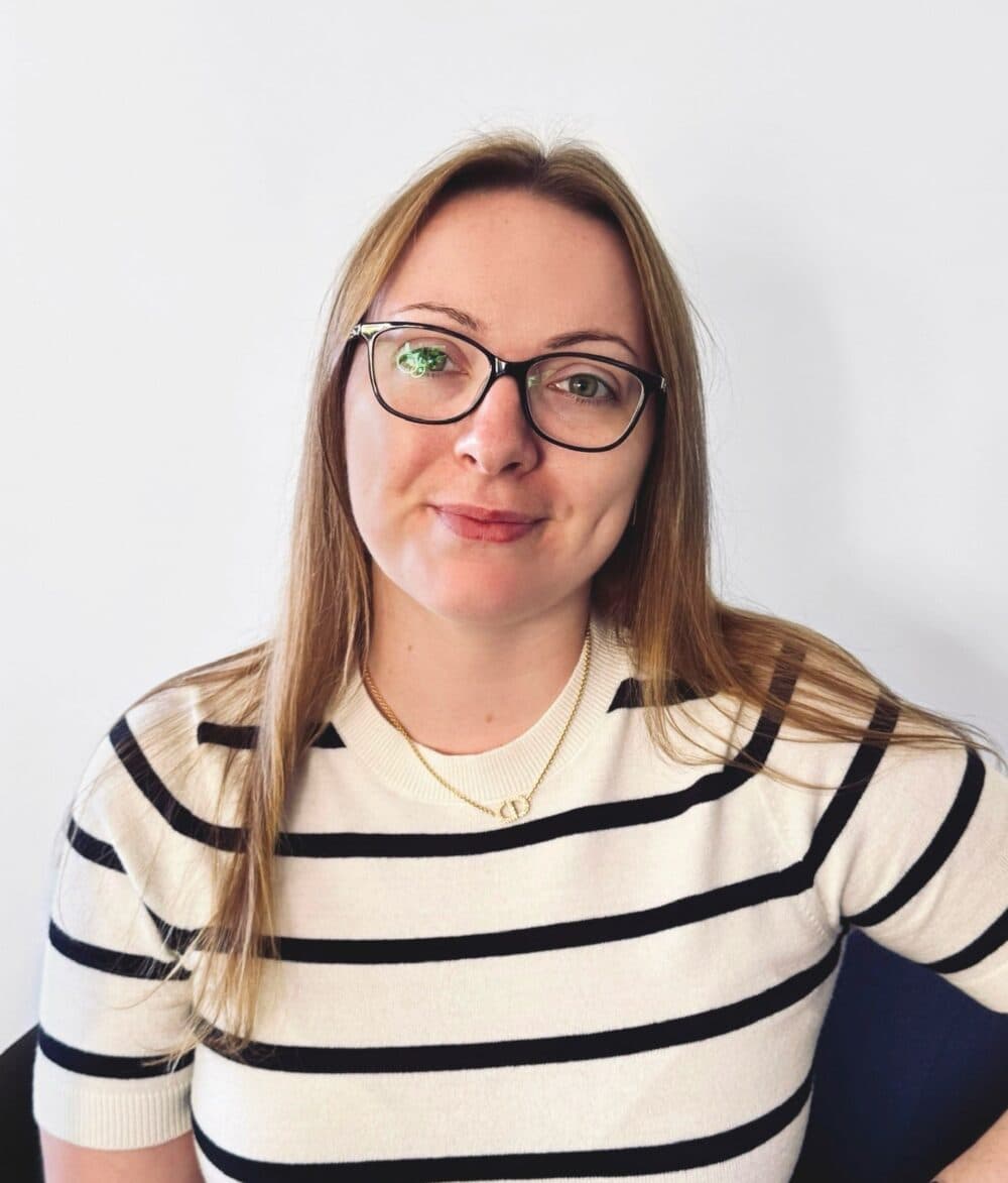 Smiling woman with glasses and long hair, wearing a striped shirt, sitting against a plain background. - Home Instead