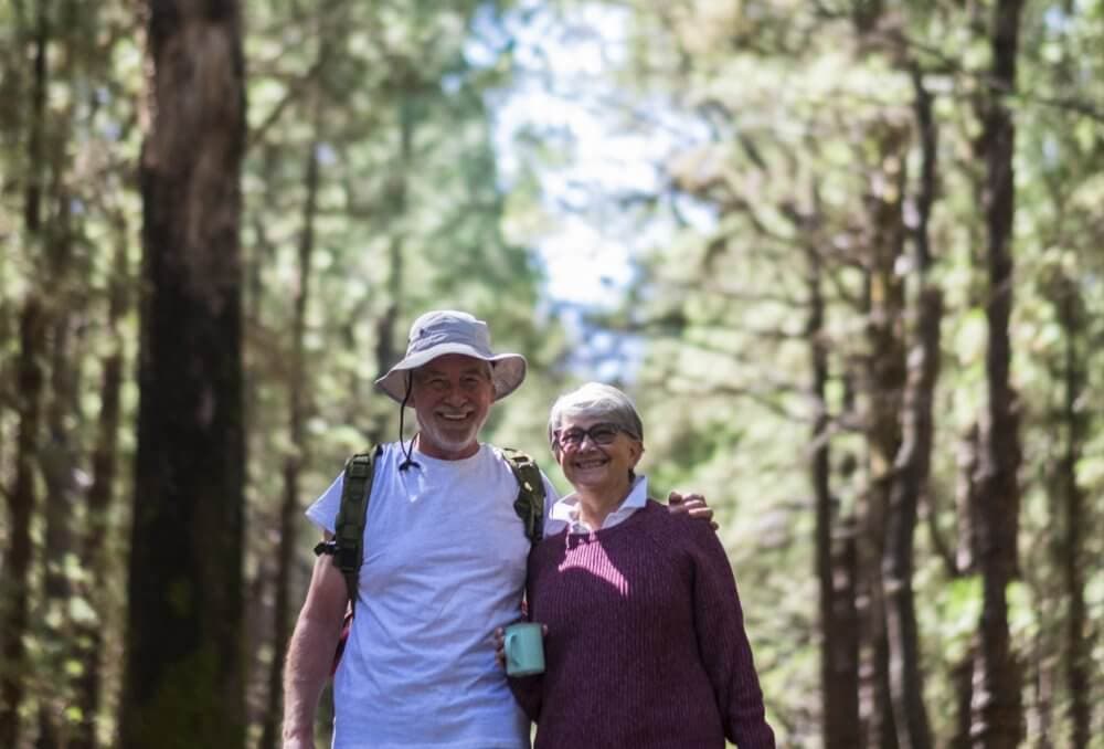 An elderly couple smiling and hugging while hiking in a forest, with trees lining the path and sunlight filtering through. - Home Instead
