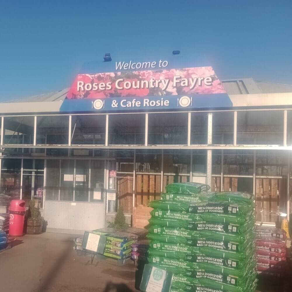 Entrance to "Roses Country Fayre & Cafe Rosie" with gardening supplies and plant sacks in front. Sign above door. - Home Instead