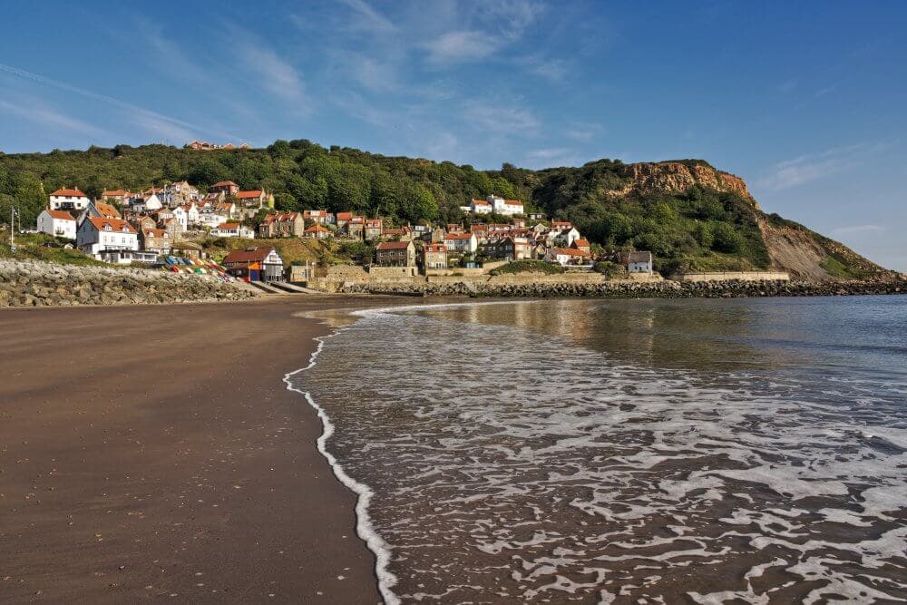 A coastal village with houses on a hillside, a sandy beach, and gentle waves under a clear blue sky. - Home Instead