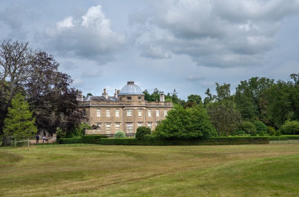 Historic mansion with a domed roof surrounded by lush gardens and trees under a partly cloudy sky. - Home Instead