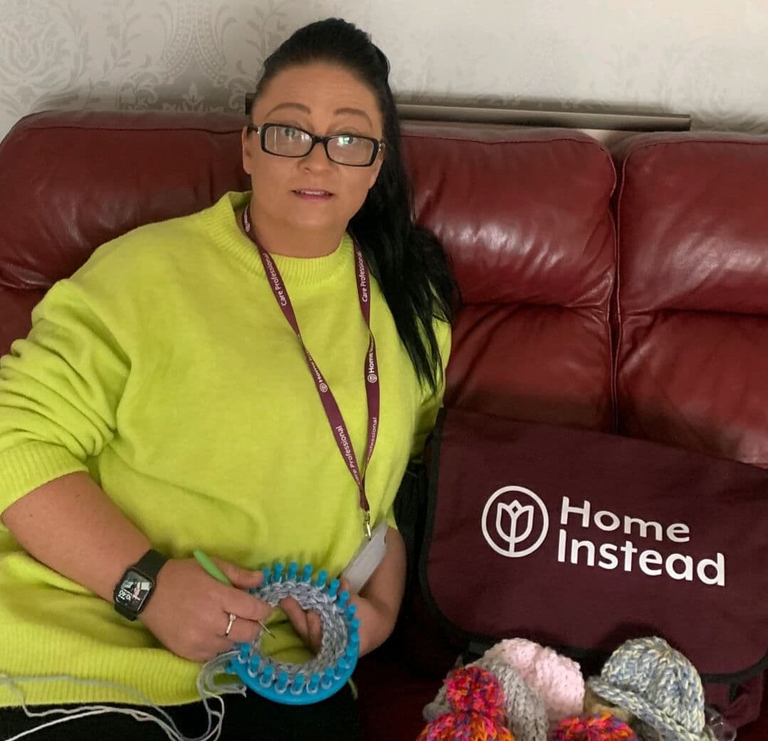 A woman with glasses knits on a loom while sitting on a red sofa, with a bag labeled "Home Instead" beside her. - Home Instead
