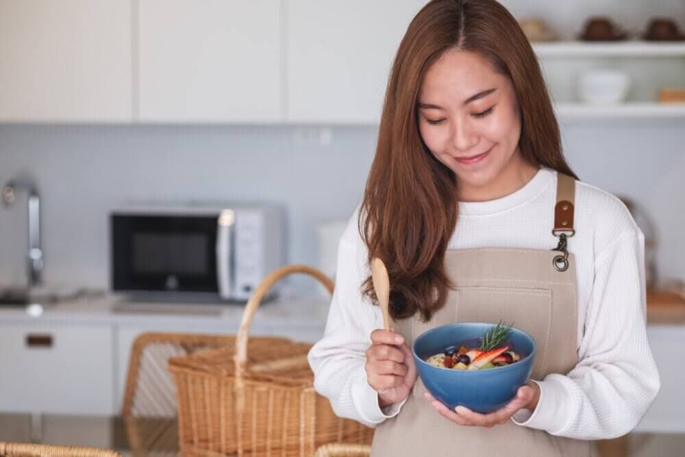 A woman in an apron holds a bowl of food in a modern kitchen, smiling and looking down at it. - Home Instead