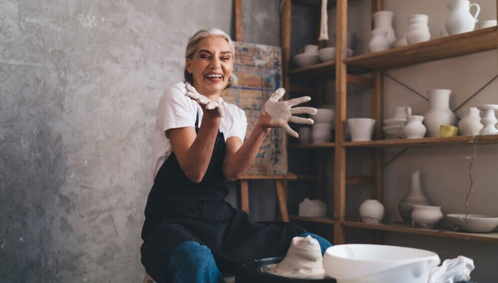 A smiling woman in an apron, covered in clay, makes pottery on a wheel in a studio with shelves of pottery behind her. - Home Instead