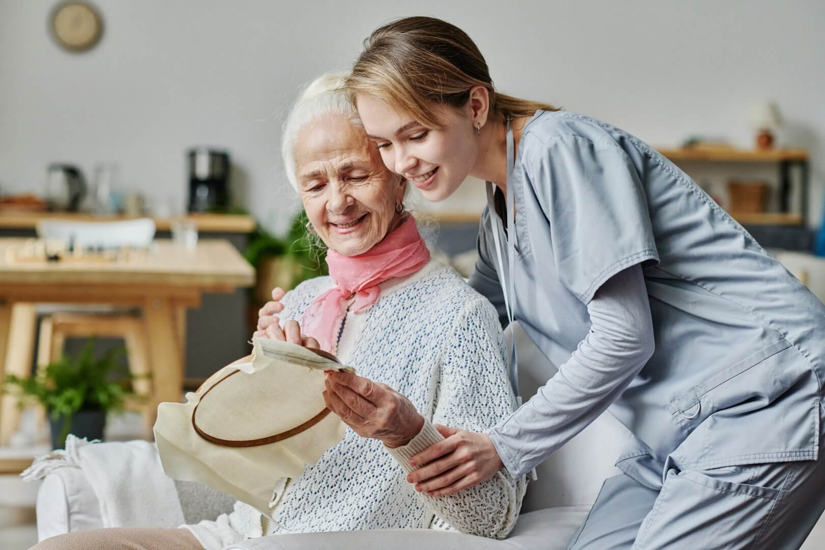 A young caregiver in scrubs assists a smiling elderly woman with embroidery in a cozy living room. - Home Instead