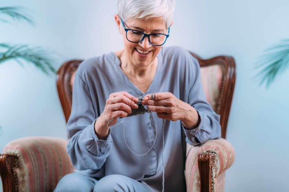 Elderly woman with short white hair knitting while sitting on an upholstered chair, smiling and focused on her work. - Home Instead