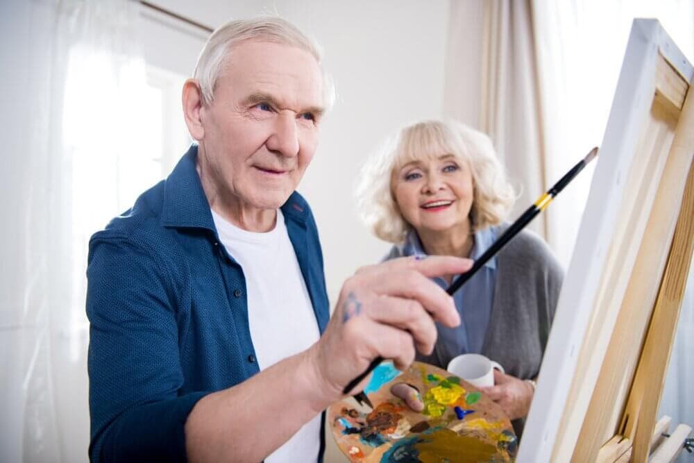 An elderly man paints on a canvas while an elderly woman smiles beside him, holding a cup. - Home Instead