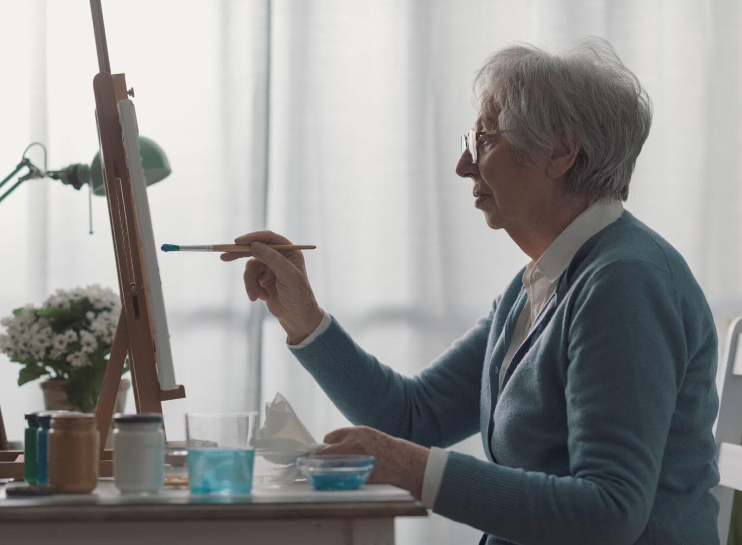 Elderly woman with grey hair painting on a canvas in a studio, with art supplies and flowers on the table. - Home Instead Bournemouth & Christchurch