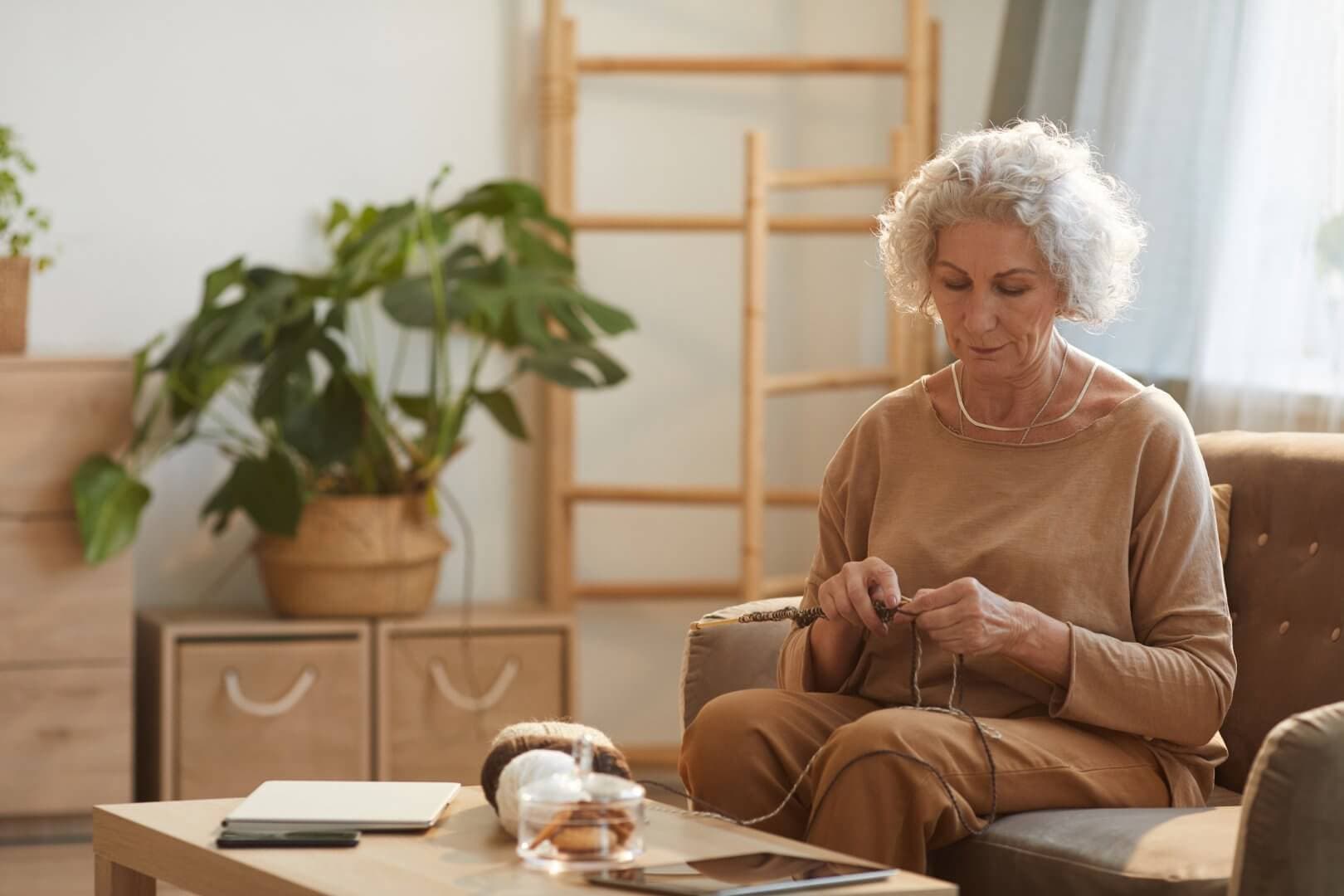 An elderly woman sits on a couch knitting, with yarn and a plant on the table, in a cozy, well-lit living room. - Home Instead