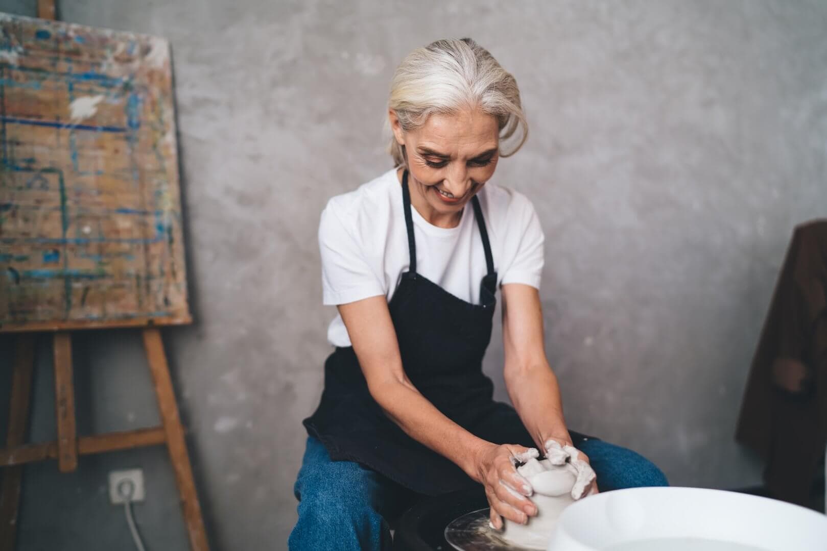 Person with white hair and blue apron focused on shaping clay on a pottery wheel in a studio. - Home Instead
