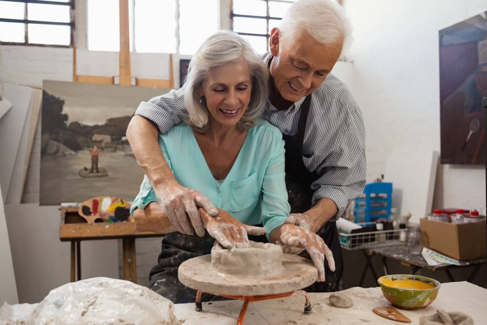 An elderly couple smiles while working together on a pottery wheel in an art studio. - Home Instead Poole