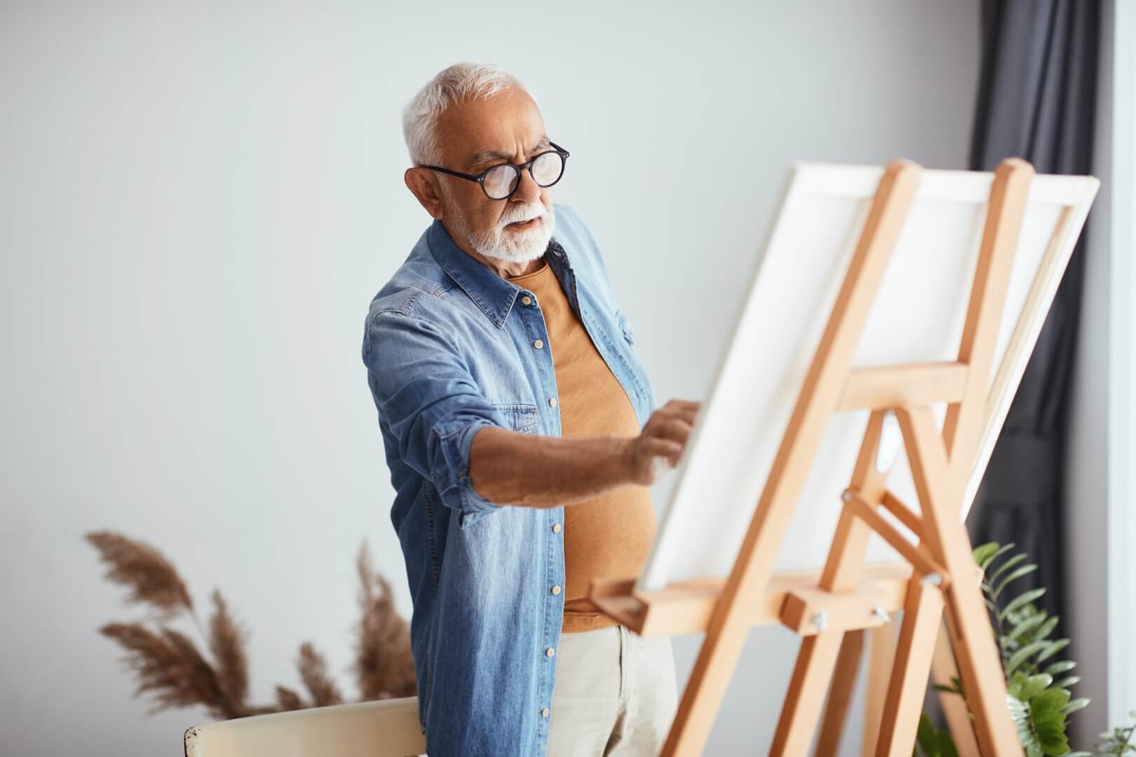 An elderly man wearing glasses and a denim shirt paints on a canvas at an easel in a bright room. - Home Instead