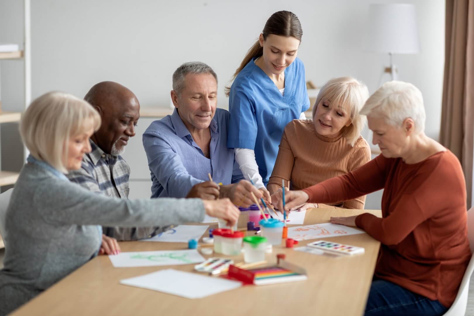 Group of diverse elderly people and a caregiver painting and drawing at a table in a bright, well-lit room. - Home Instead