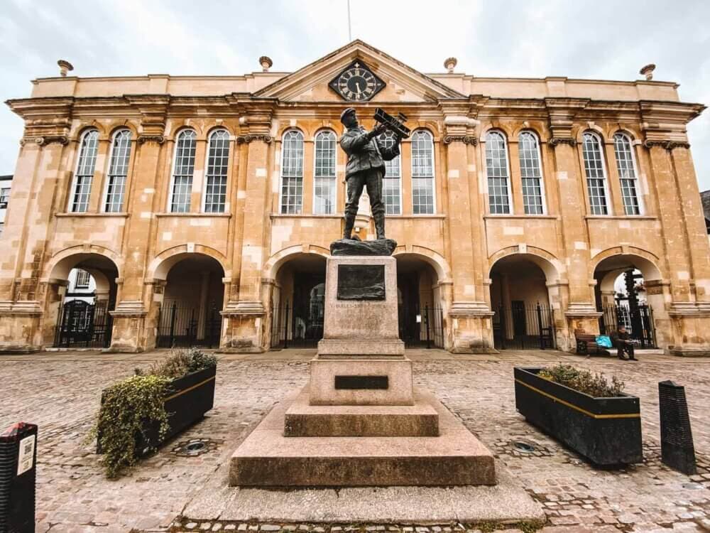 Statue of a man holding a rifle in front of a historic building with arched windows and clock above the entrance. - Home Instead