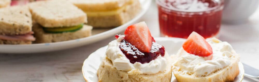 Scones with cream and jam, topped with strawberries, are in the foreground, with sandwiches and a glass of jam in the back. - Home Instead