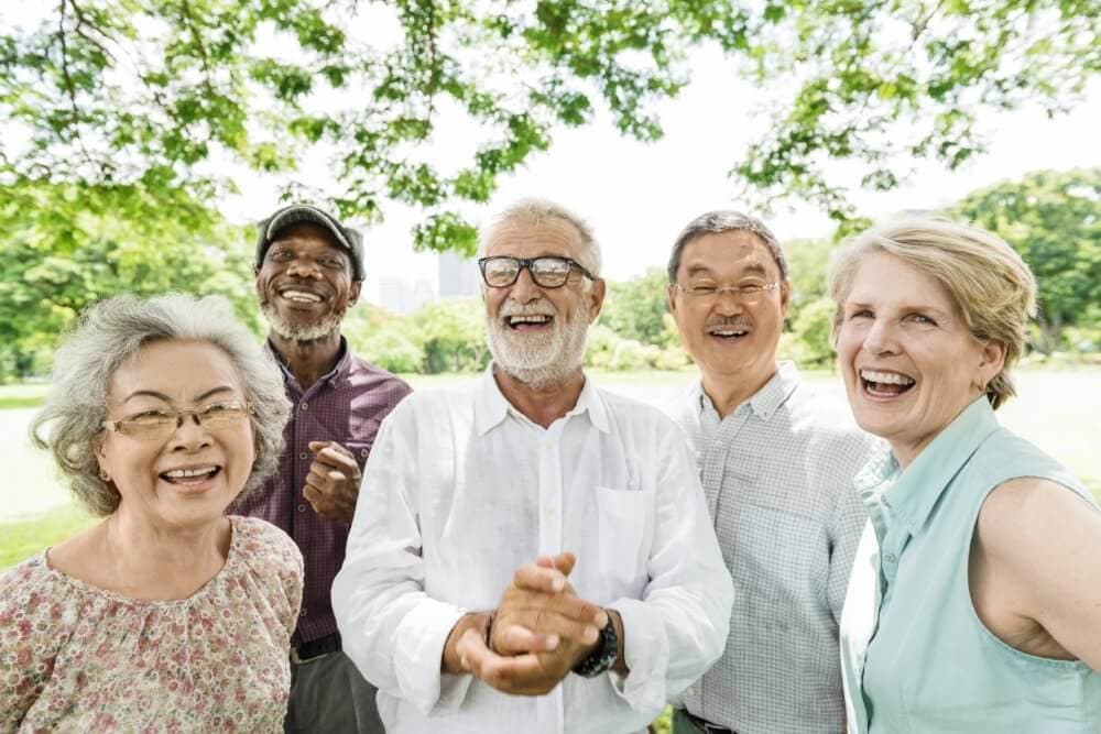 Five smiling older adults standing outdoors beneath trees, enjoying a sunny day together. - Home Instead
