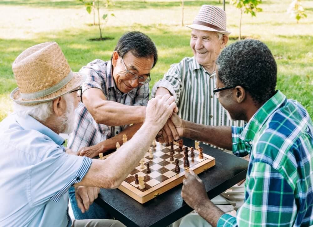 Four elderly men enjoying a game of chess outdoors, all smiling and holding hands over the chessboard. - Home Instead