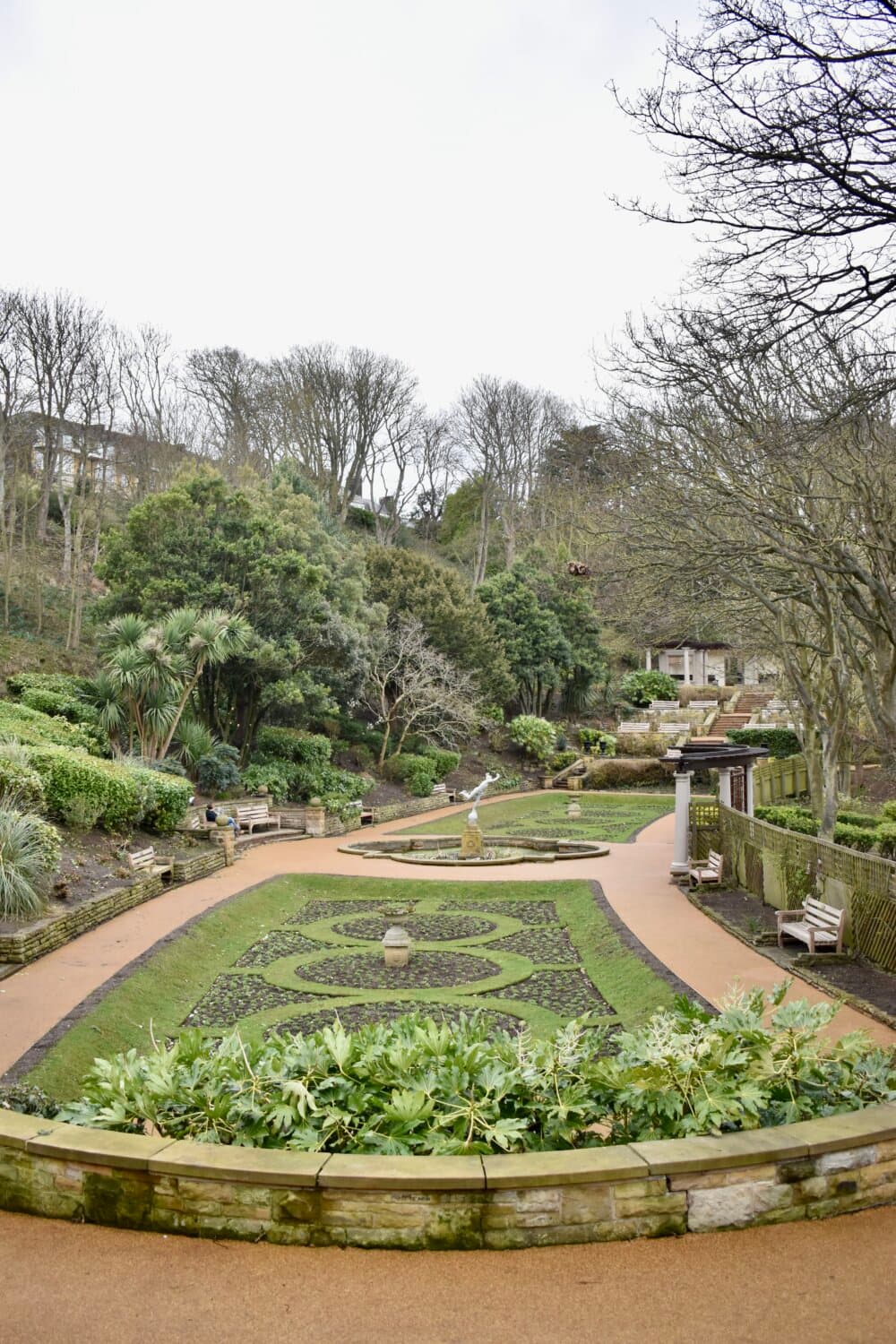 A landscaped garden with hedges, benches, pathways, and trees, with a statue in the center, on a cloudy day. - Home Instead