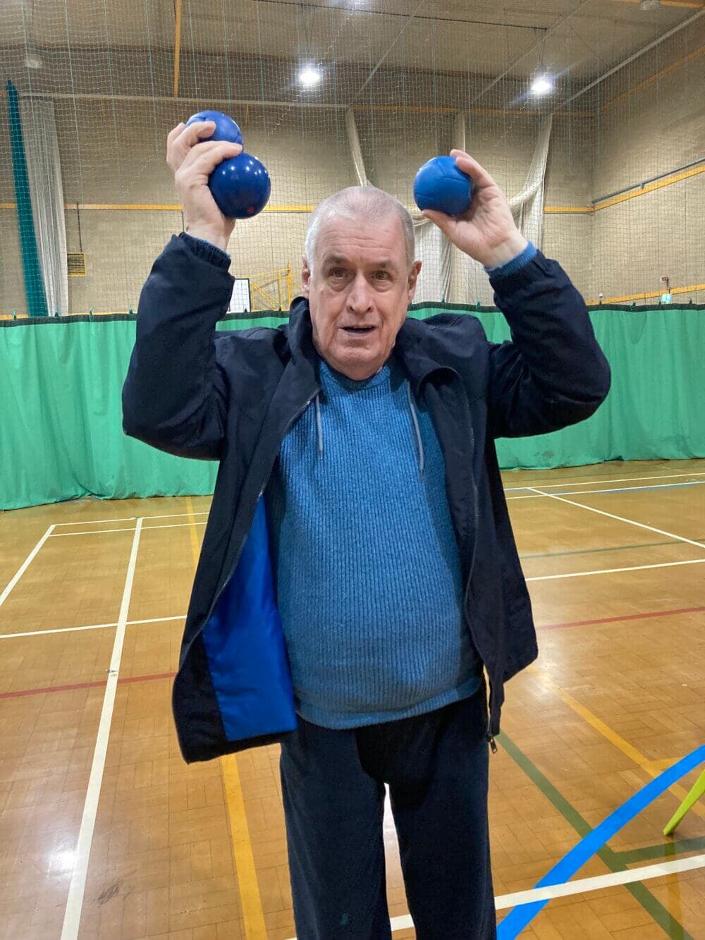 A man in a blue sweater and black jacket holds up two blue balls in a gymnasium with green netted walls. - Home Instead
