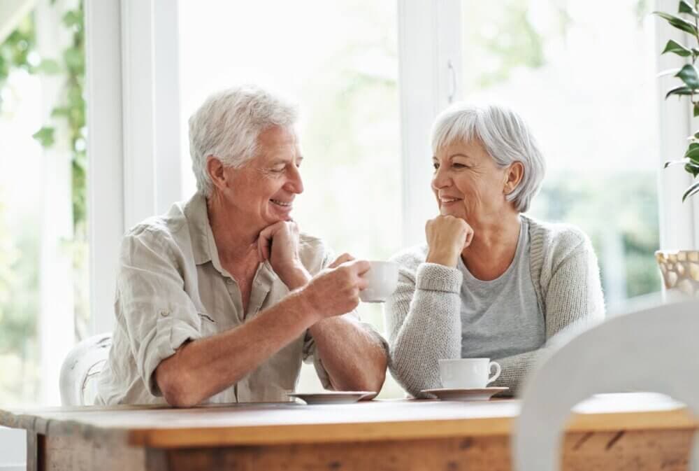Elderly man and woman sitting at a wooden table, smiling at each other while drinking coffee or tea. - Home Instead Poole