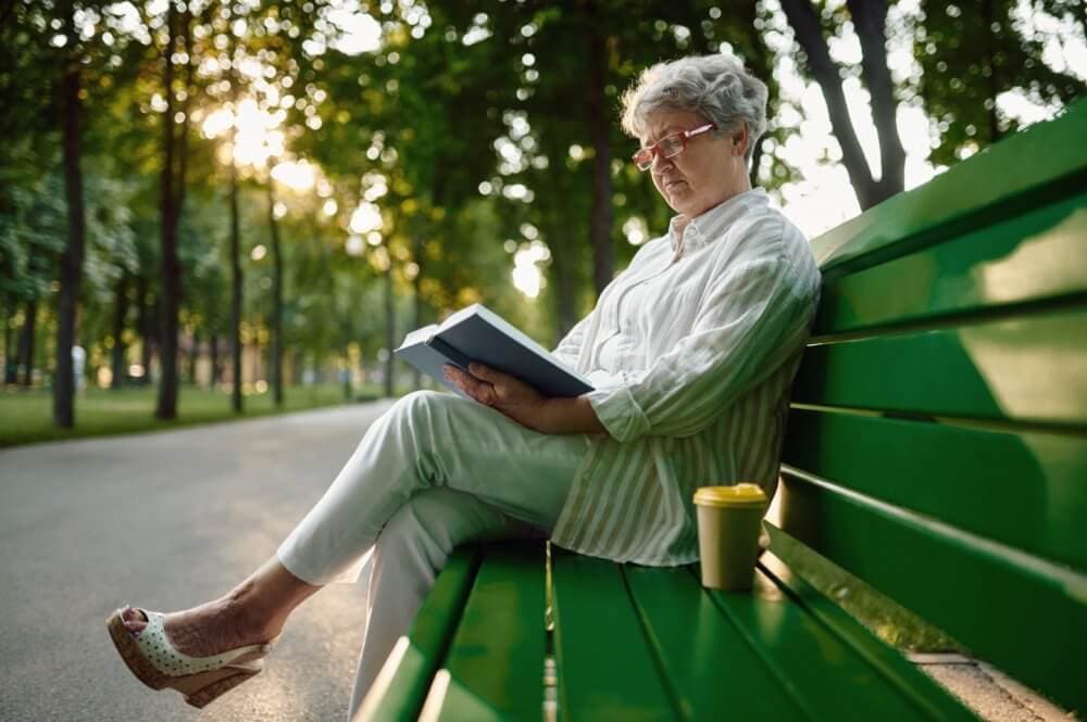 Elderly person reading a book on a green bench in a park with a takeaway coffee cup beside them. - Home Instead Southampton
